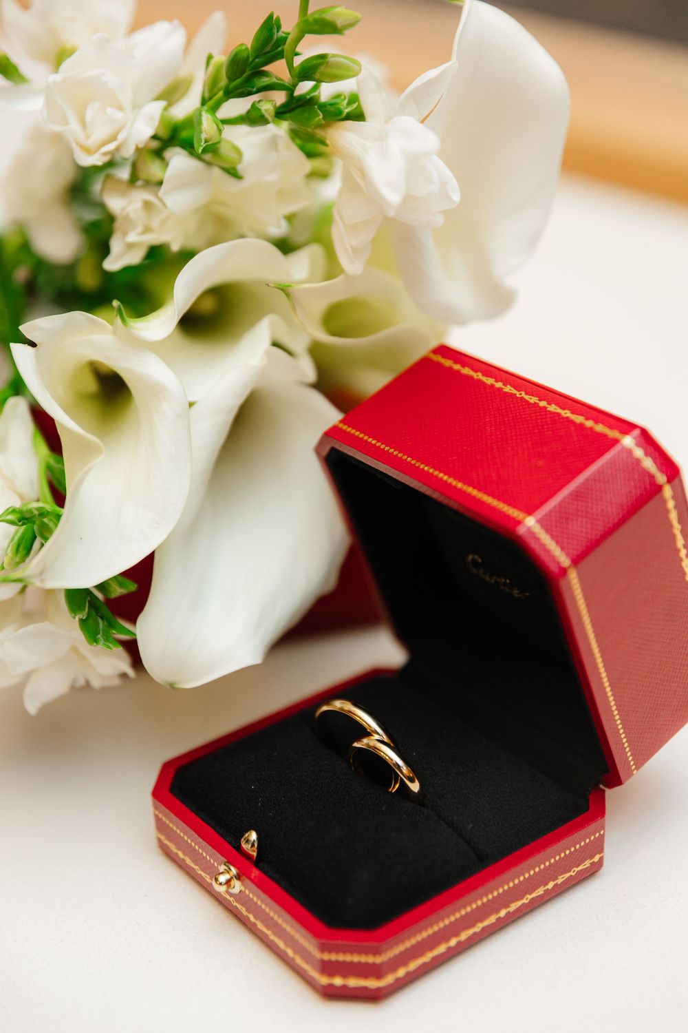 Wedding ring in red velvet box with elegant white calla lilies and greenery bouquet in the background.
