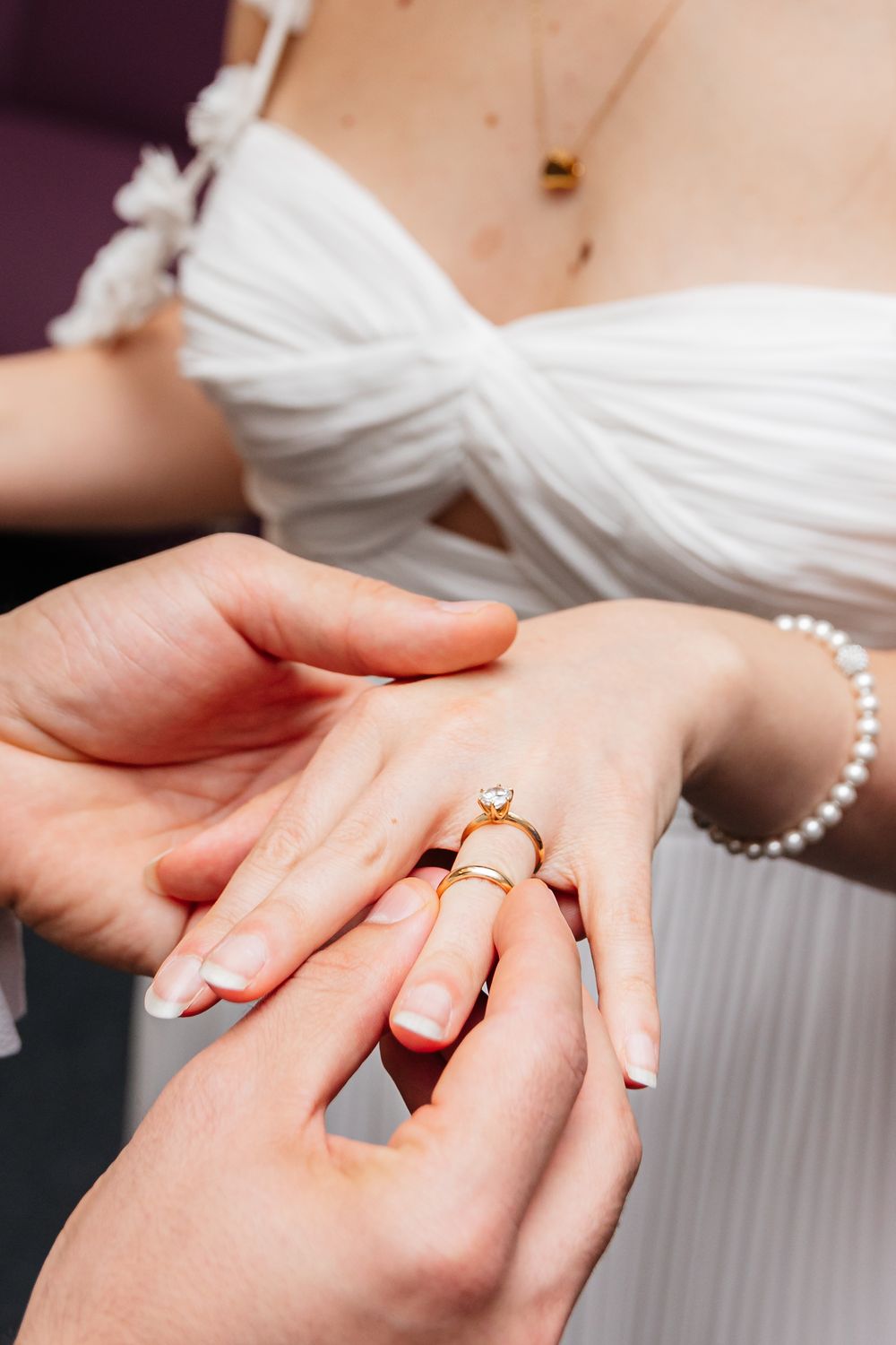 A close-up shot of hands placing an engagement ring during a wedding ceremony, with a white dress visible in background.