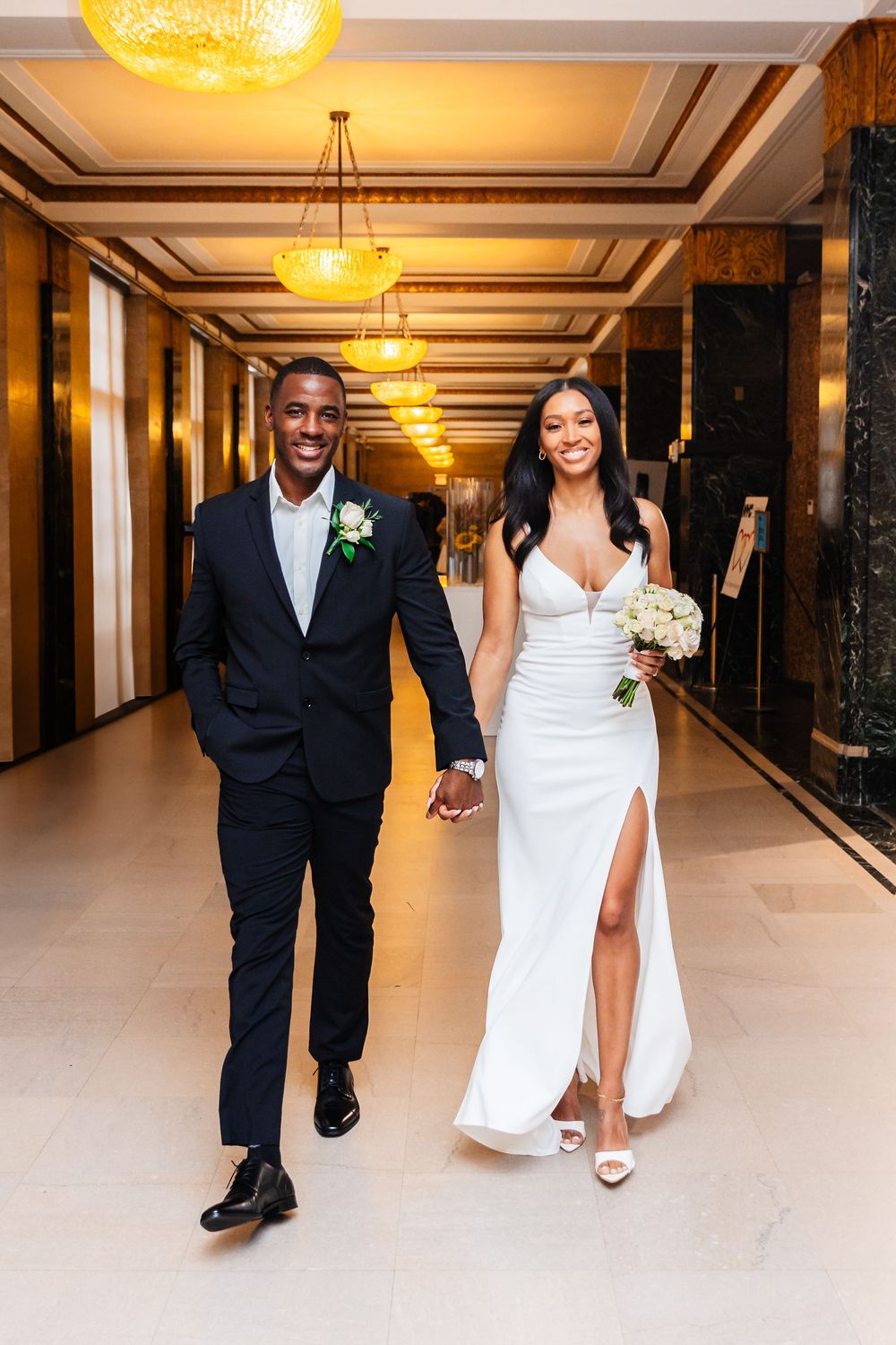 A happy couple walks hand in hand down an elegant hotel hallway with gold lighting and marble columns.