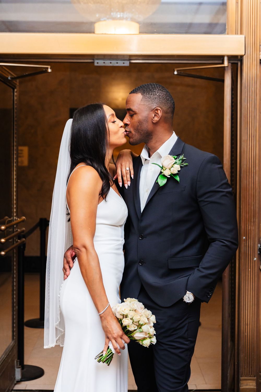 A couple shares an intimate kiss at their wedding, wearing elegant white gown and black suit with floral boutonniere.