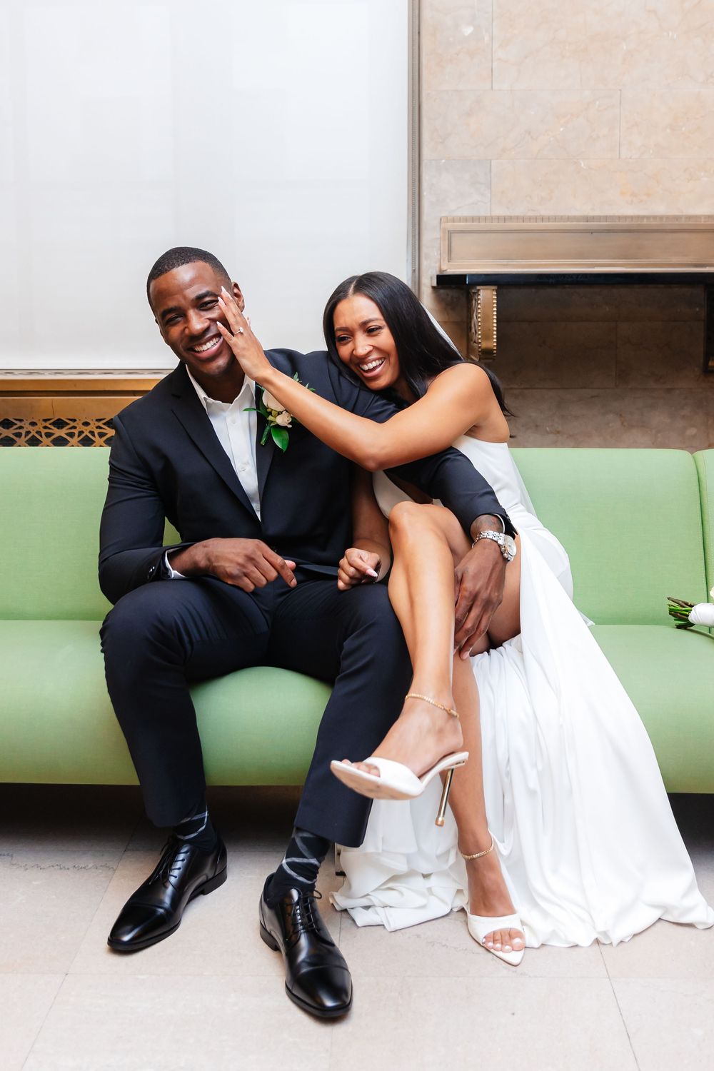 A happy couple in wedding attire sits together on a green couch, sharing joyful moments in a photo series.