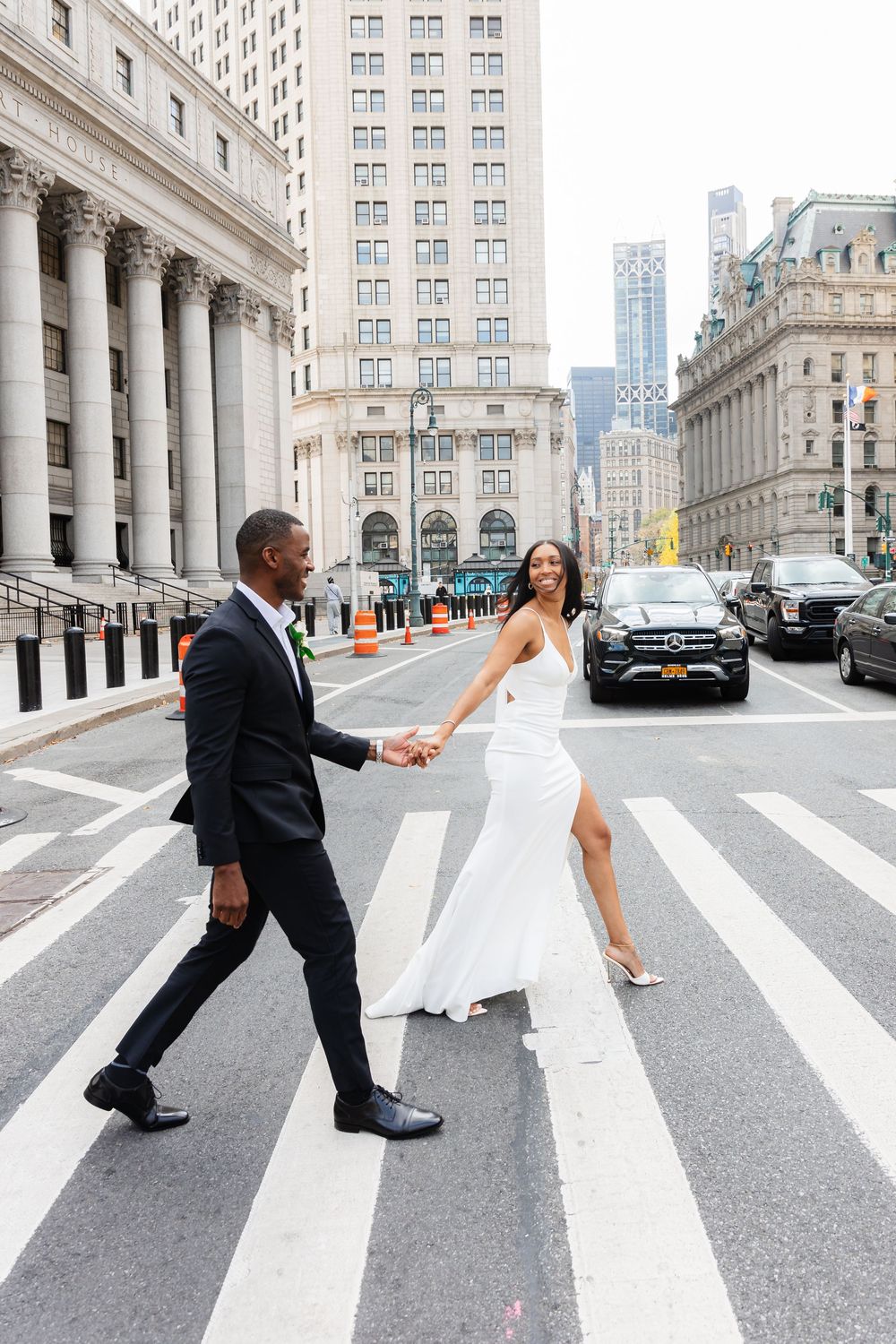 A wedding couple recreates the iconic Abbey Road album cover pose on a city crosswalk with tall buildings behind them.
