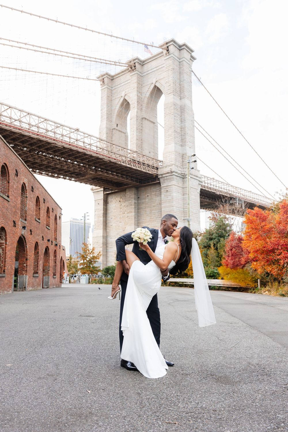 A romantic wedding couple shares a dip kiss beneath the iconic Brooklyn Bridge with autumn foliage in the background.
