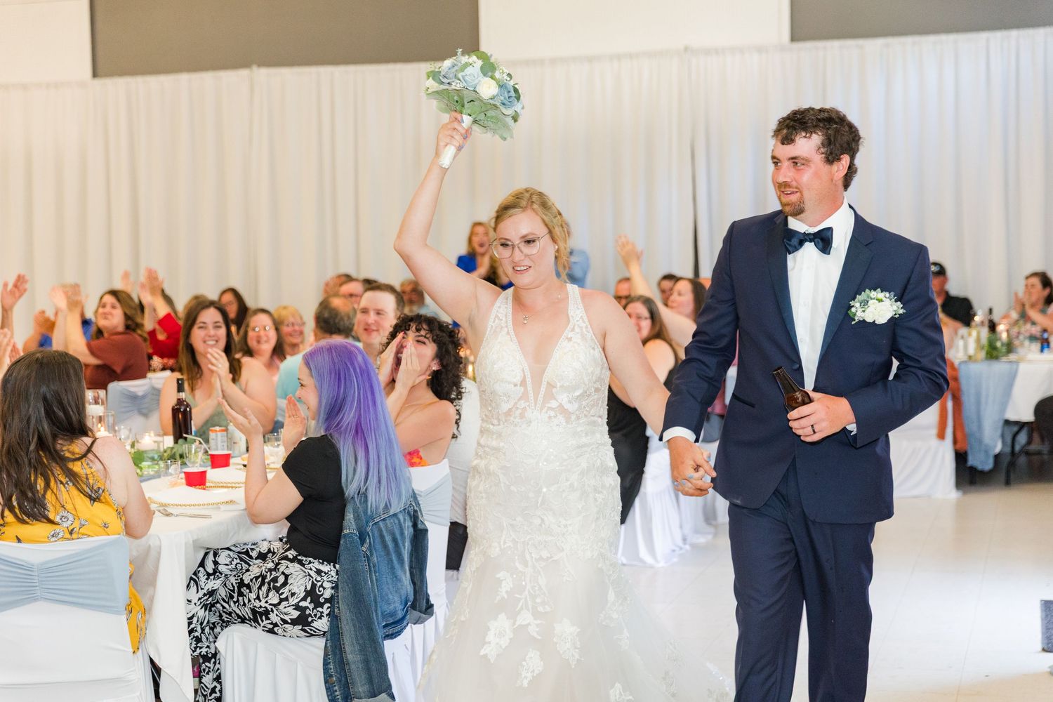 Newlyweds celebrate and walk through reception hall while wedding guests cheer and applaud.
