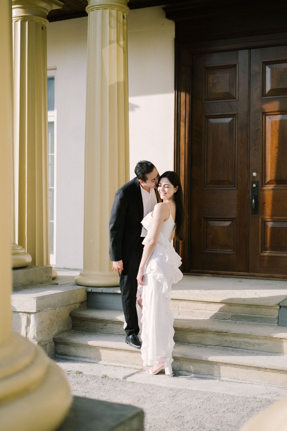 A couple in formal wedding attire stands on stone steps with elegant yellow columns and dark wooden doors in the background.