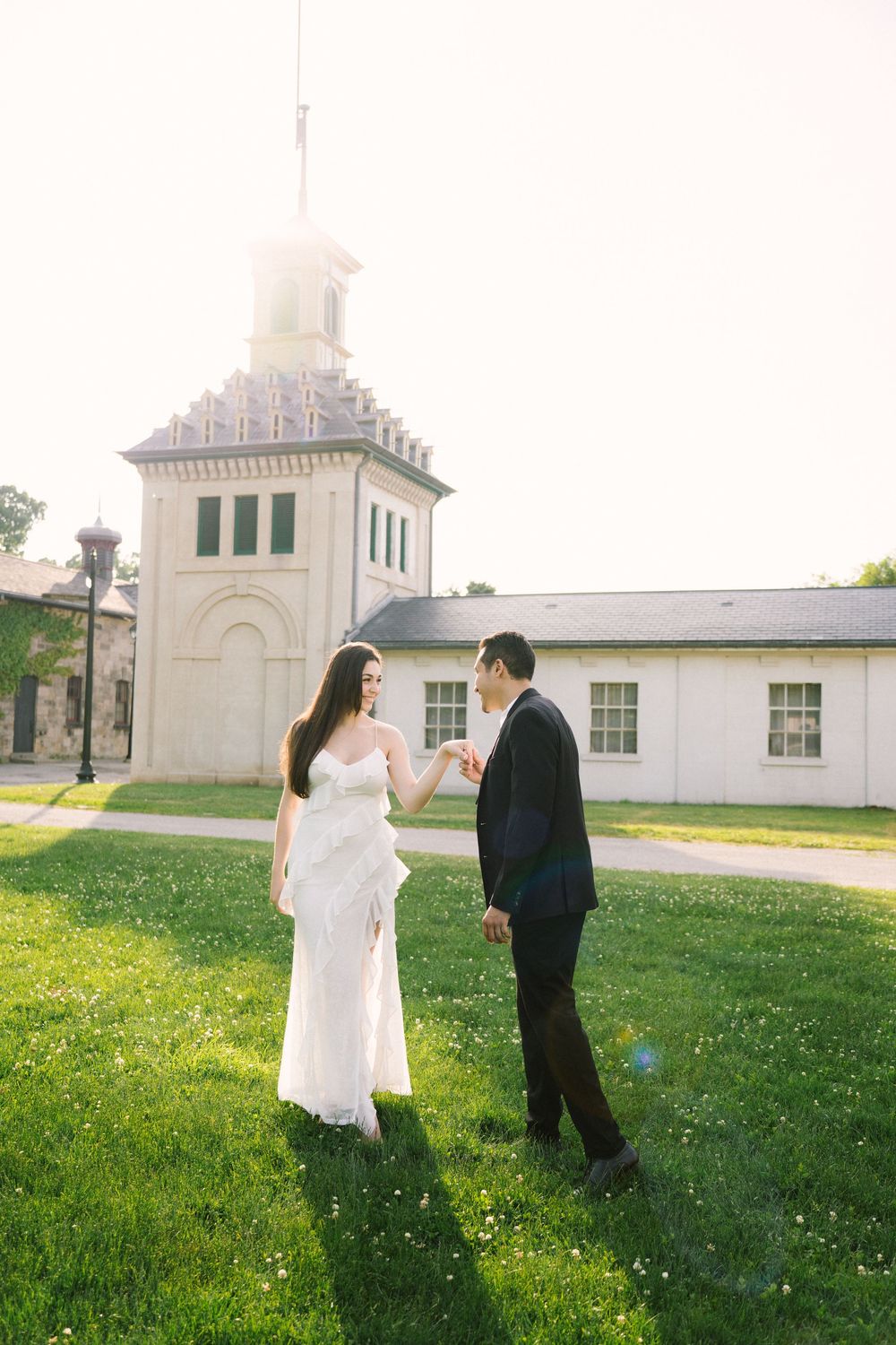 A couple holding hands walks together on grassy field near white church buildings on a sunny spring day.