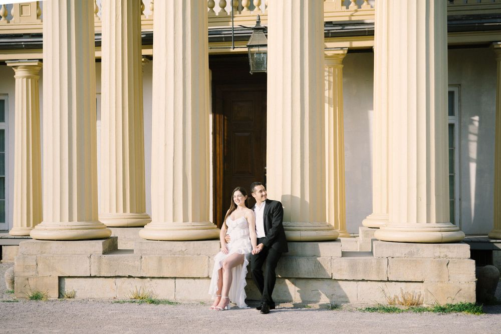 A couple stands between elegant white classical columns of a historic building in soft natural light.