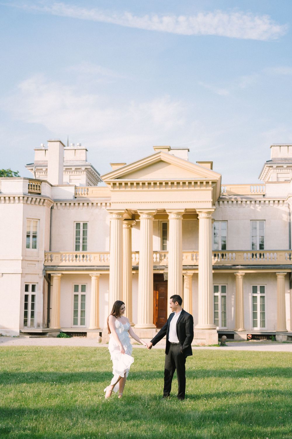 Two people stand in front of a grand neoclassical mansion with white columns and elegant architecture on a sunny day.