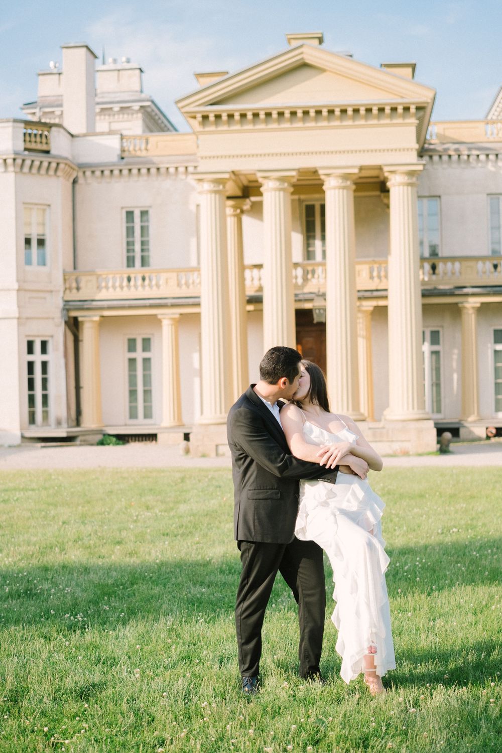 A romantic couple dances and embraces on a grassy lawn in front of an elegant neoclassical mansion with white columns.