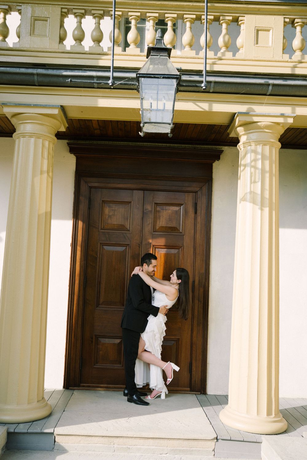 Romantic couple embraces in front of grand wooden door with classic columns and lantern overhead.