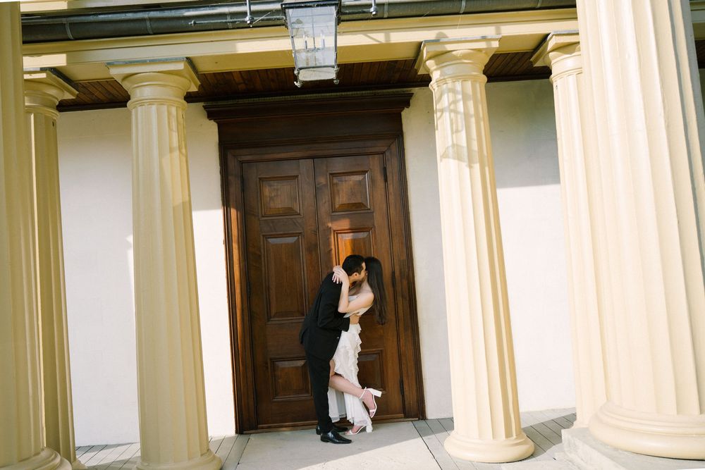 A couple embraces in a romantic pose against a wooden door framed by classic white columns.