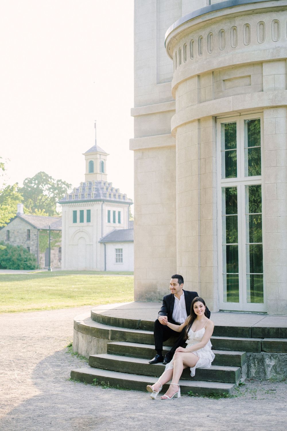 Romantic couple sits together on steps of a grand, white columned building while gazing at each other in the sunlight.