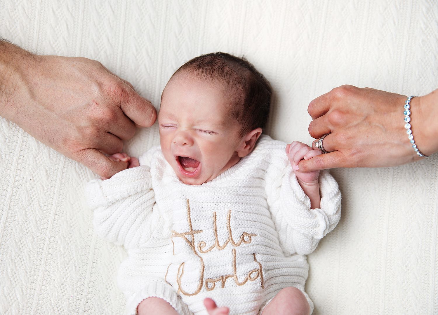 A newborn baby yawning while wearing a white 'Hello World' onesie, with adult hands on either side. During a newborn session in Bexley