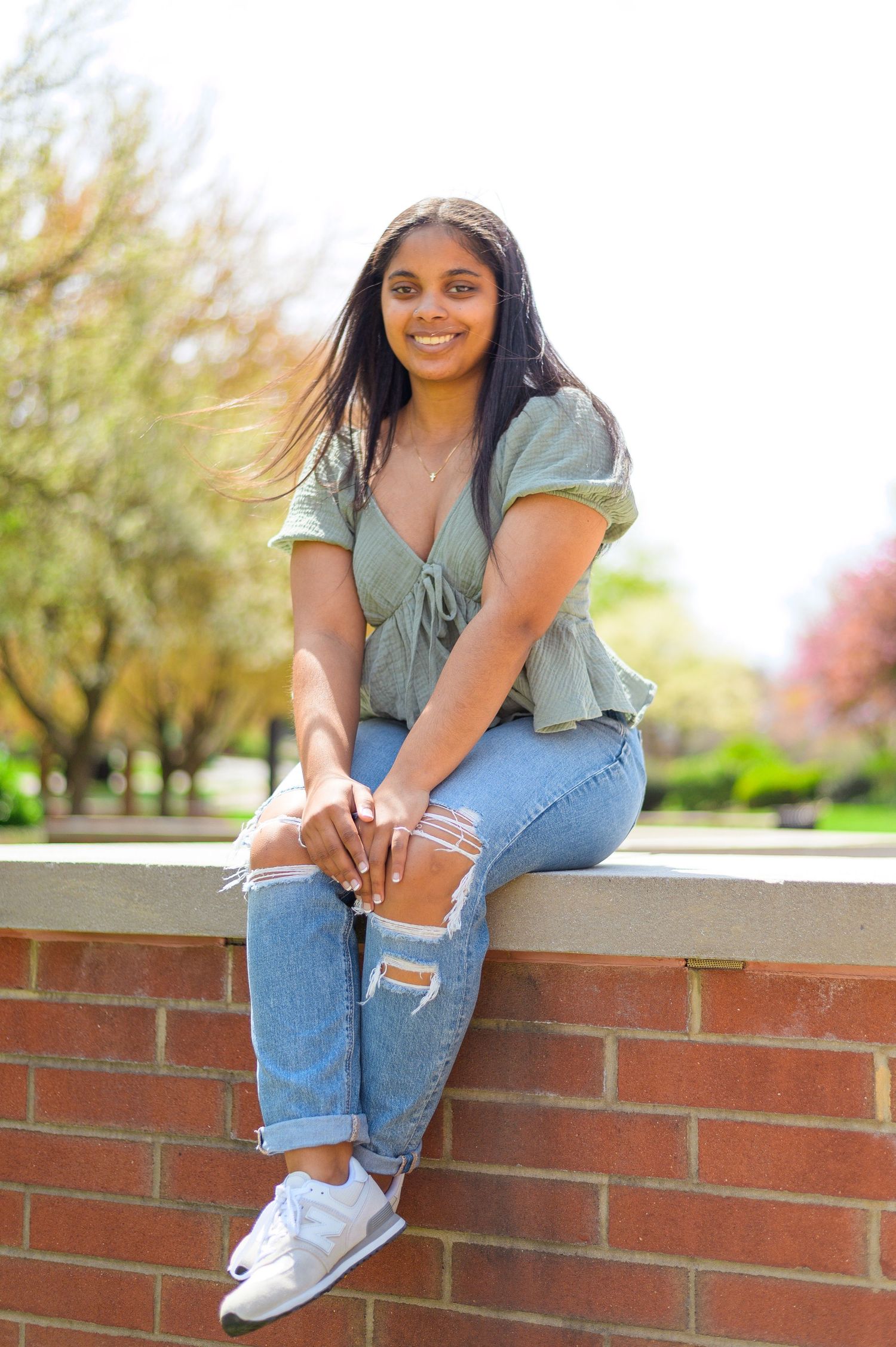 A student in casual attire sits on a brick wall during a sunny day on campus.