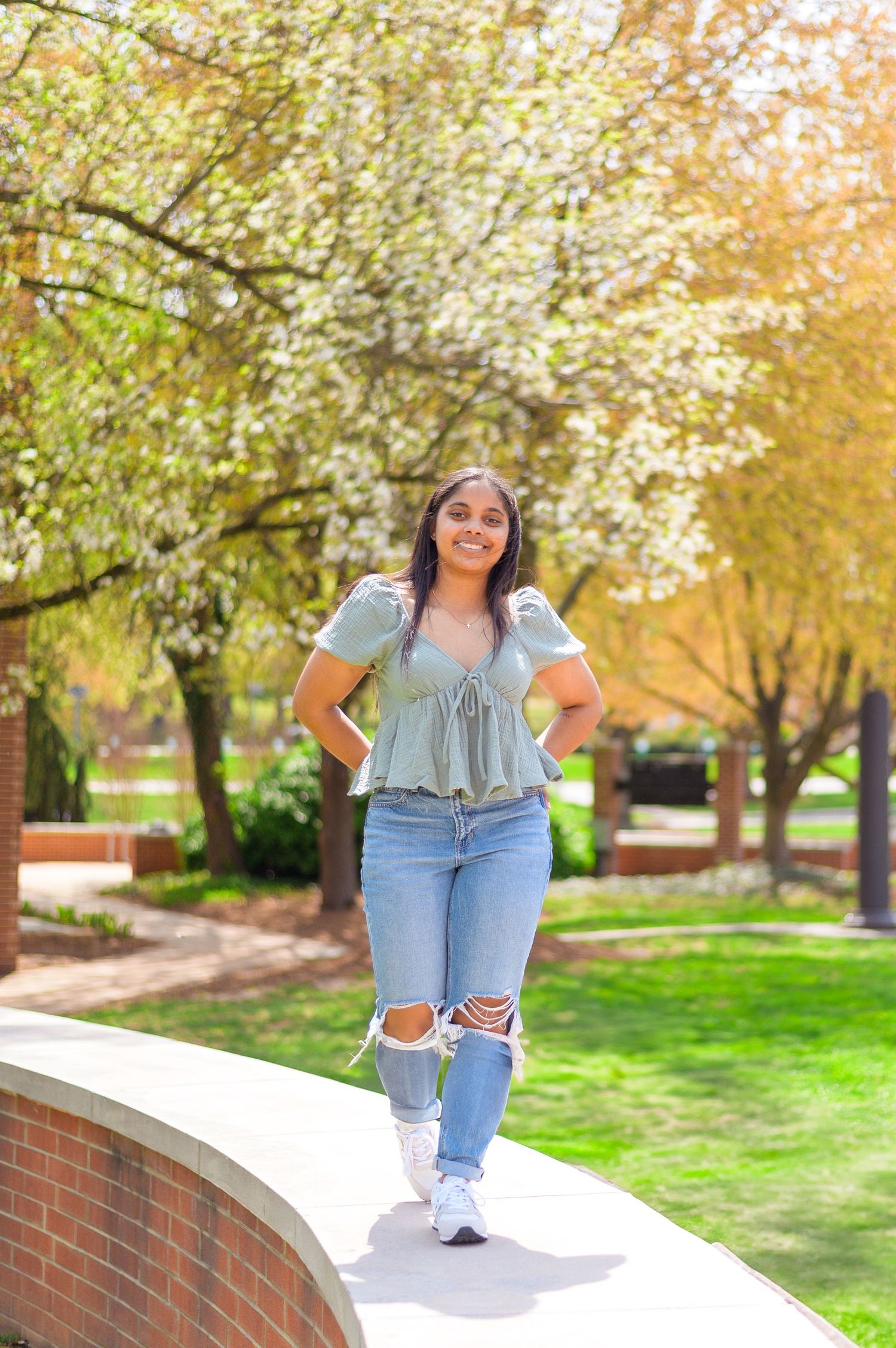 A student poses on a curved brick wall with blooming spring trees in the background.