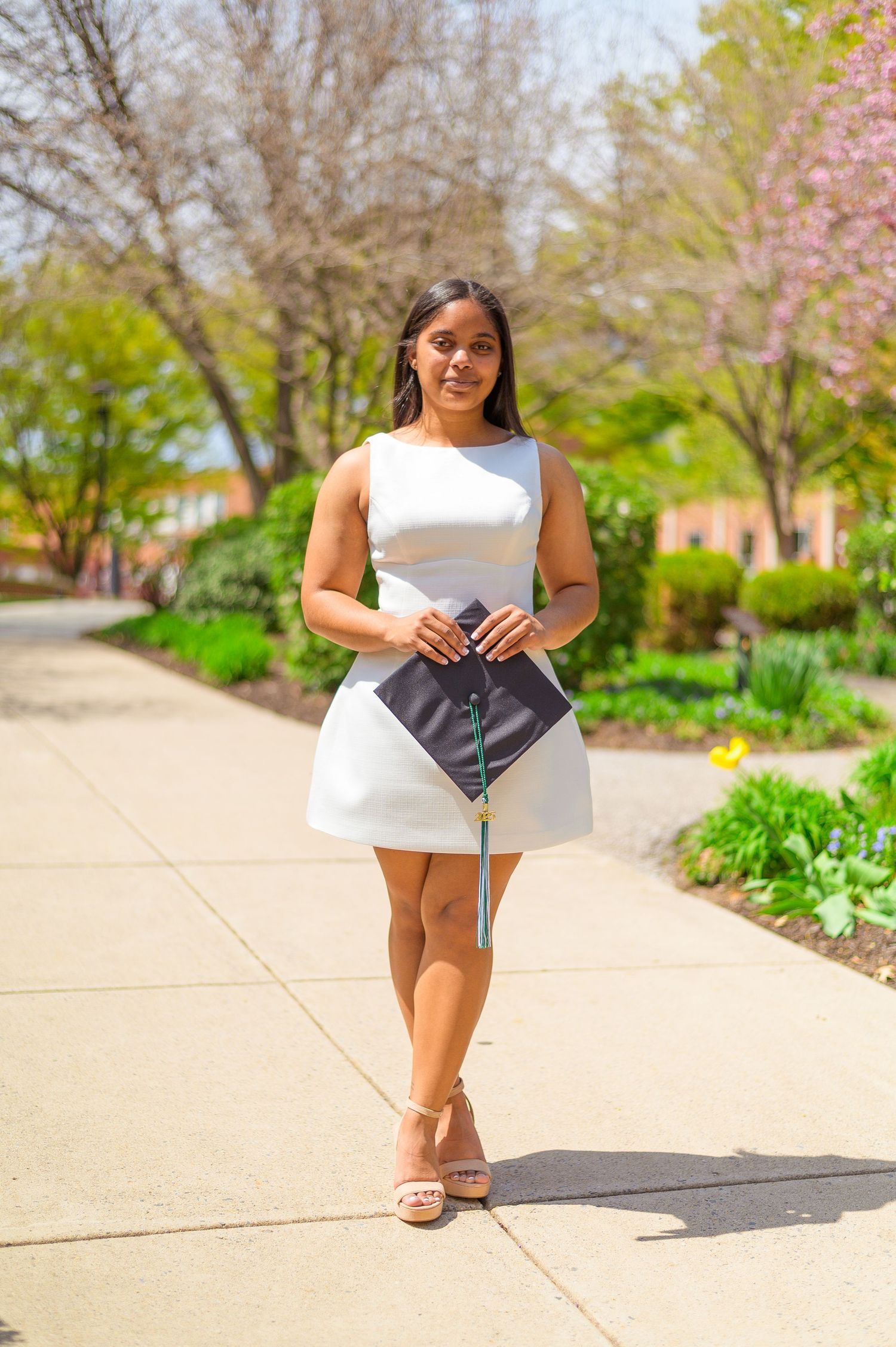 A graduate poses in a white dress holding a diploma on a sunny campus walkway.