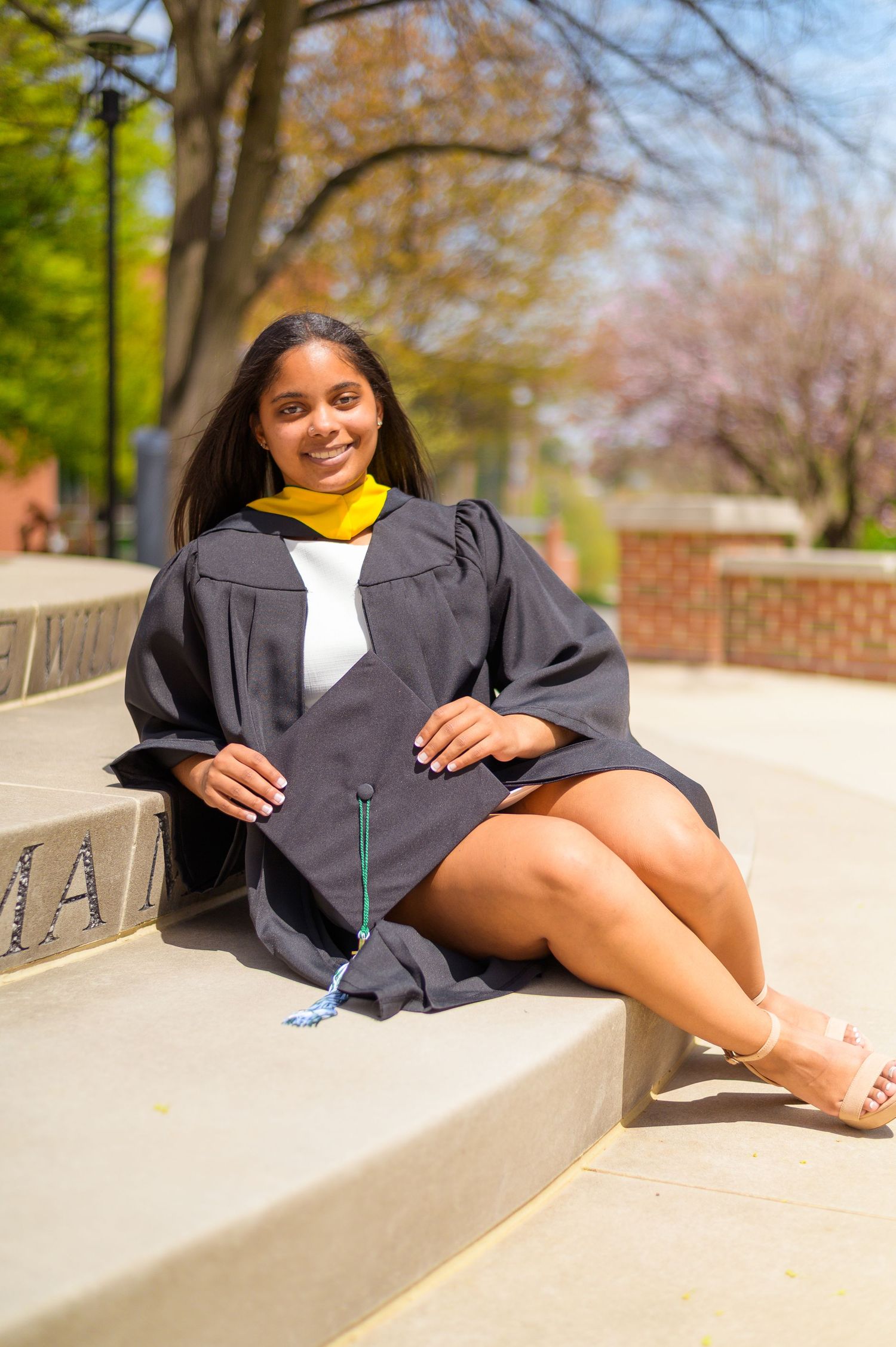 A graduate in academic regalia relaxes on concrete steps during a sunny graduation photoshoot.