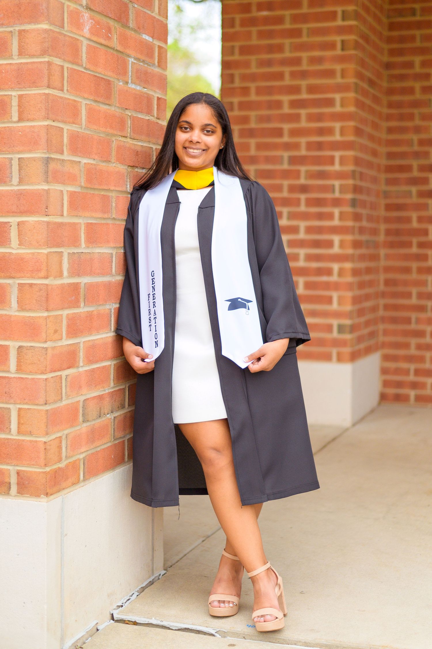 A graduate in academic regalia poses against red brick columns wearing a yellow stole.