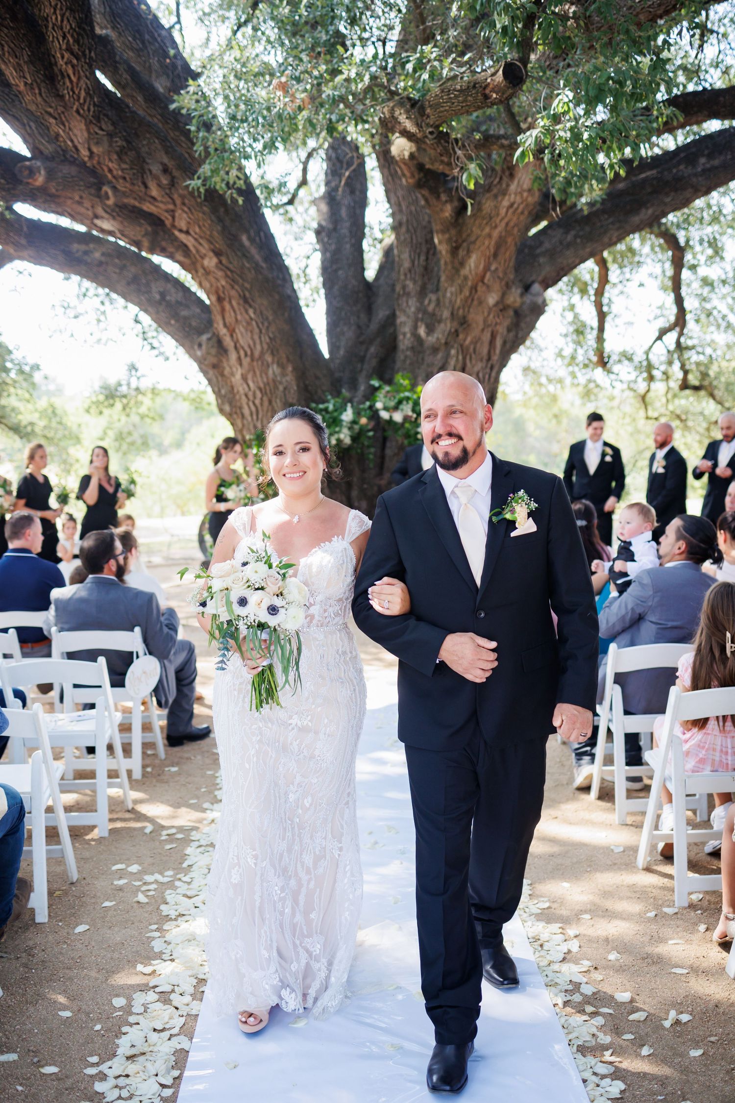 An outdoor wedding ceremony setup under large trees with white chairs arranged in rows and couples walking down the aisle.