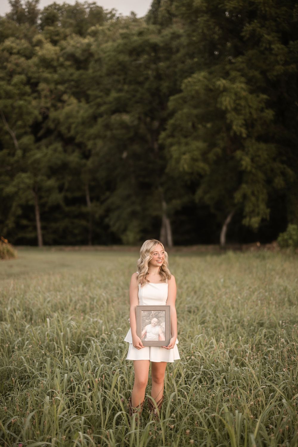 A figure in a white summer dress stands in a sunlit meadow surrounded by tall trees during golden hour.