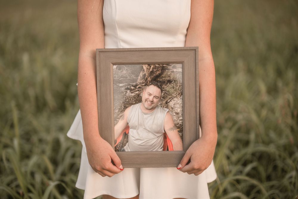 Person in white dress holds framed portrait photograph against grassy outdoor backdrop in golden sunlight.