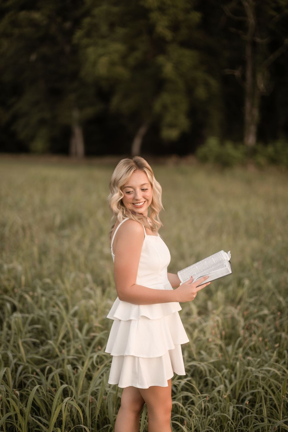 A toddler in a white dress holds a book while playing in a tall grass field on a sunny day.