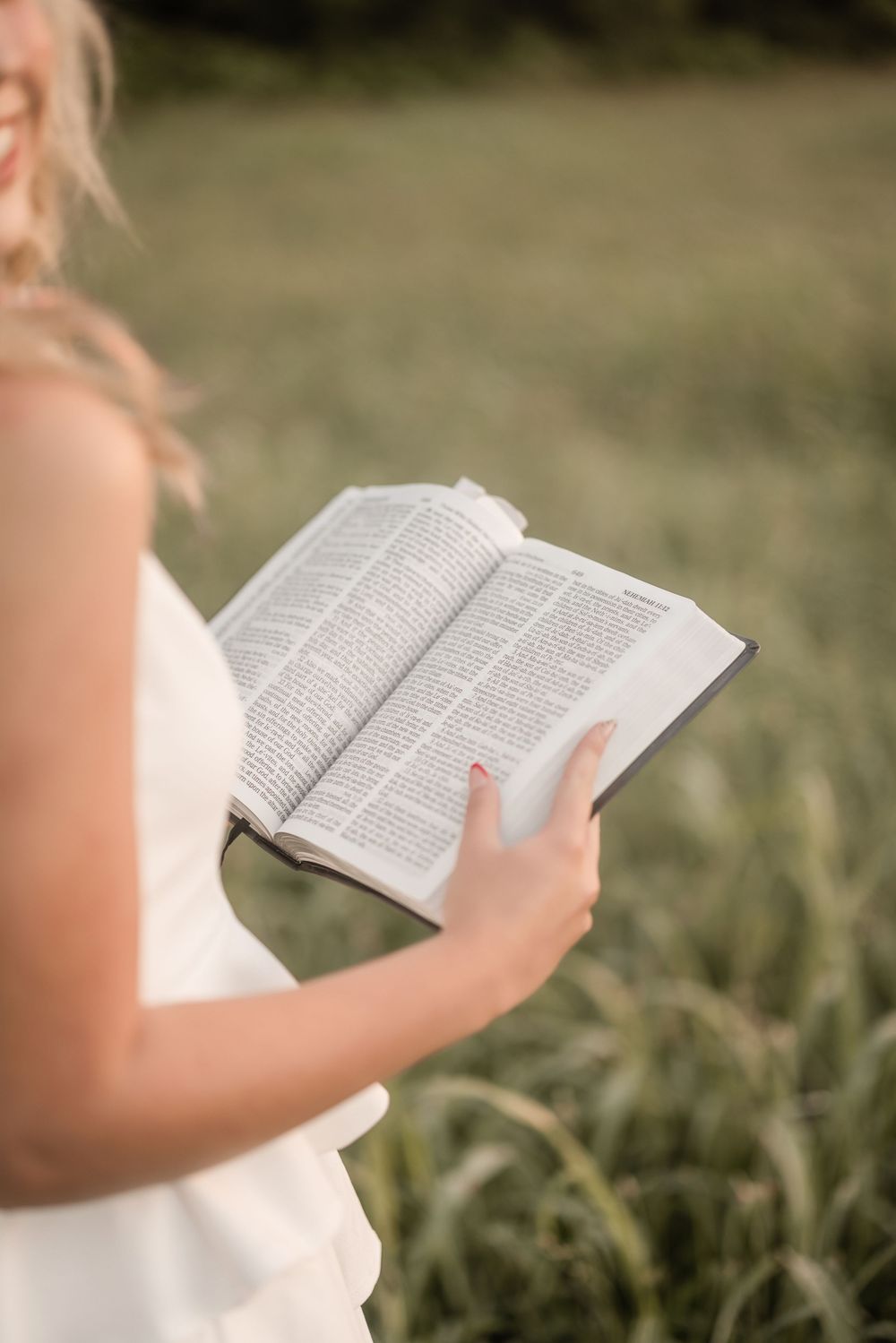 Hands holding an open book in white dress against blurred natural outdoor background.