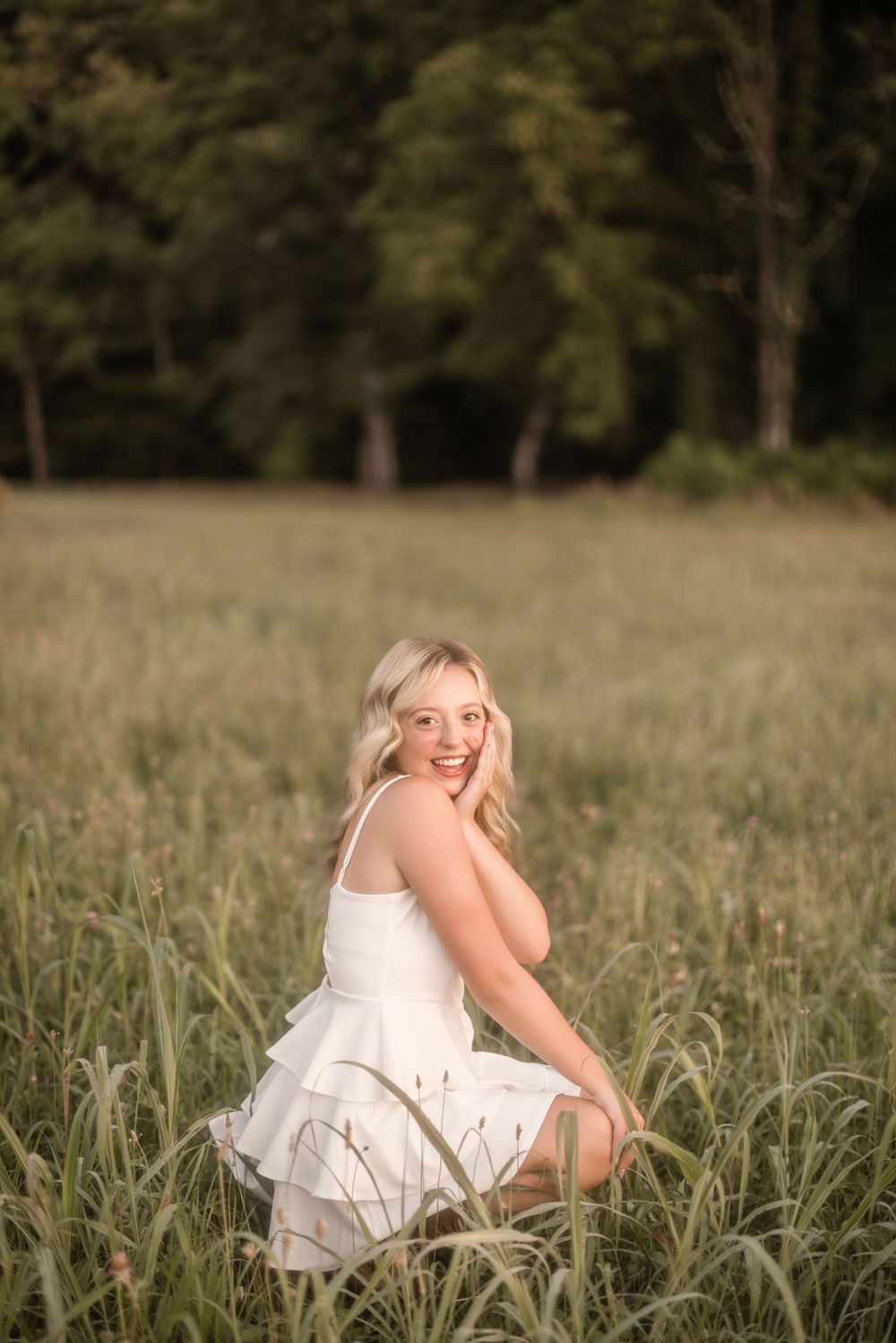 A person in a white dress sits gracefully in a sunlit meadow surrounded by tall grass and trees.