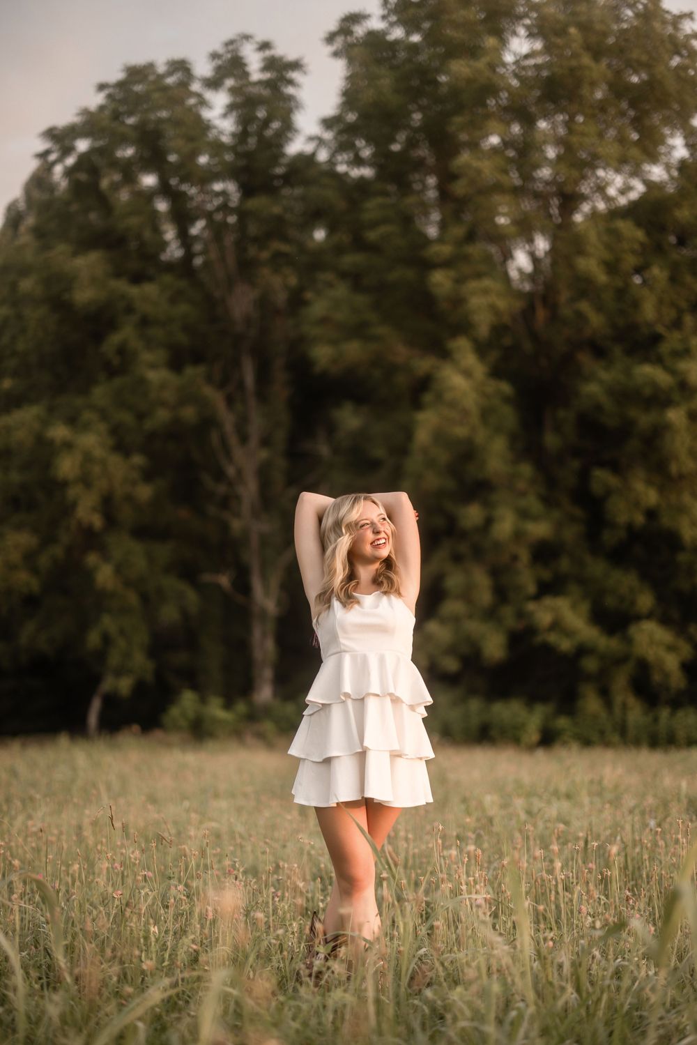 Someone in a white tiered sundress stands in a sunlit meadow with trees in the background at golden hour.
