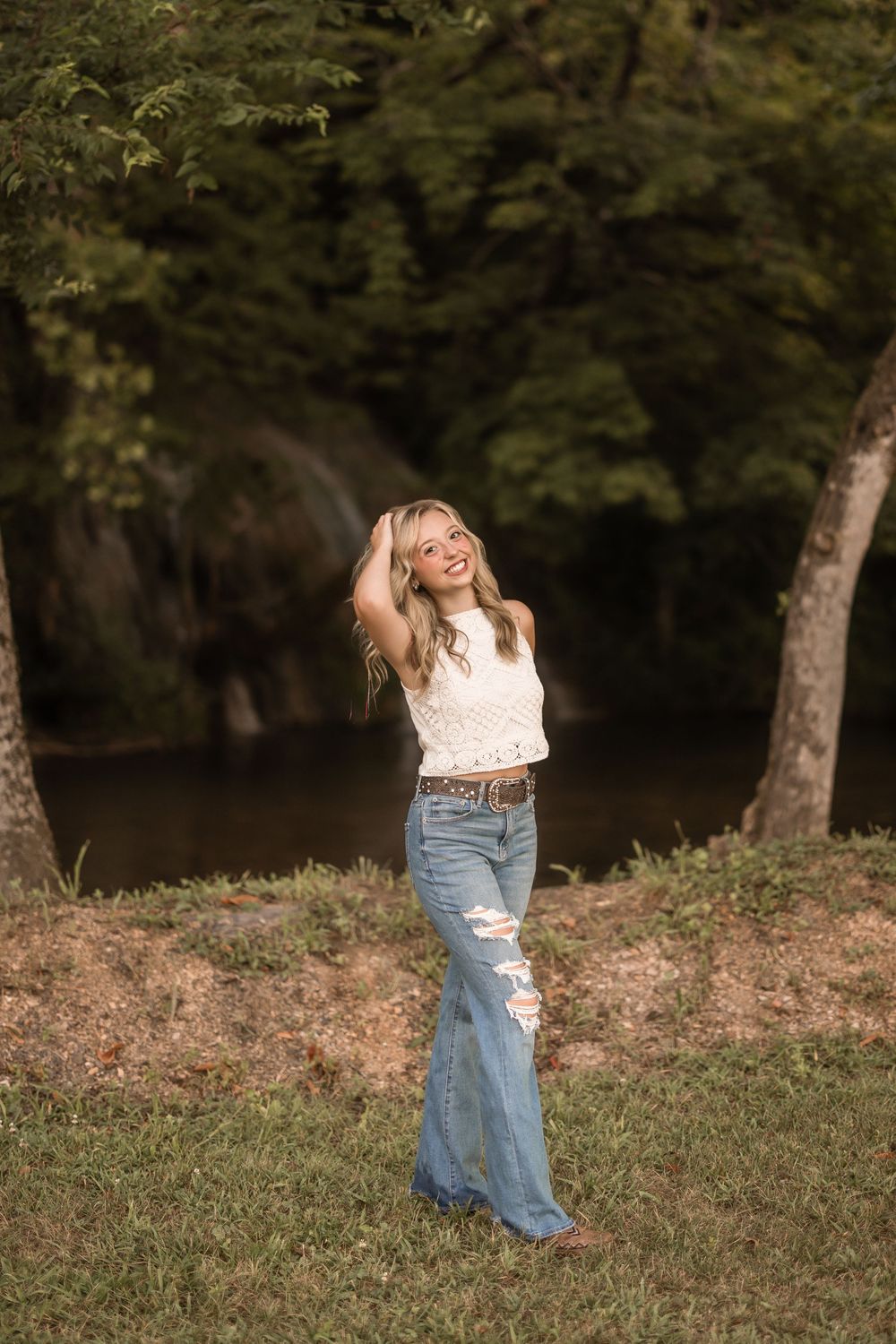 Person in denim skirt twirls joyfully on grassy hillside surrounded by trees on a sunny day.