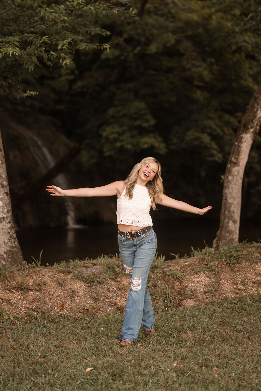 A joyful moment captured in nature showing someone in ripped jeans and white top with arms outstretched near a waterfall.