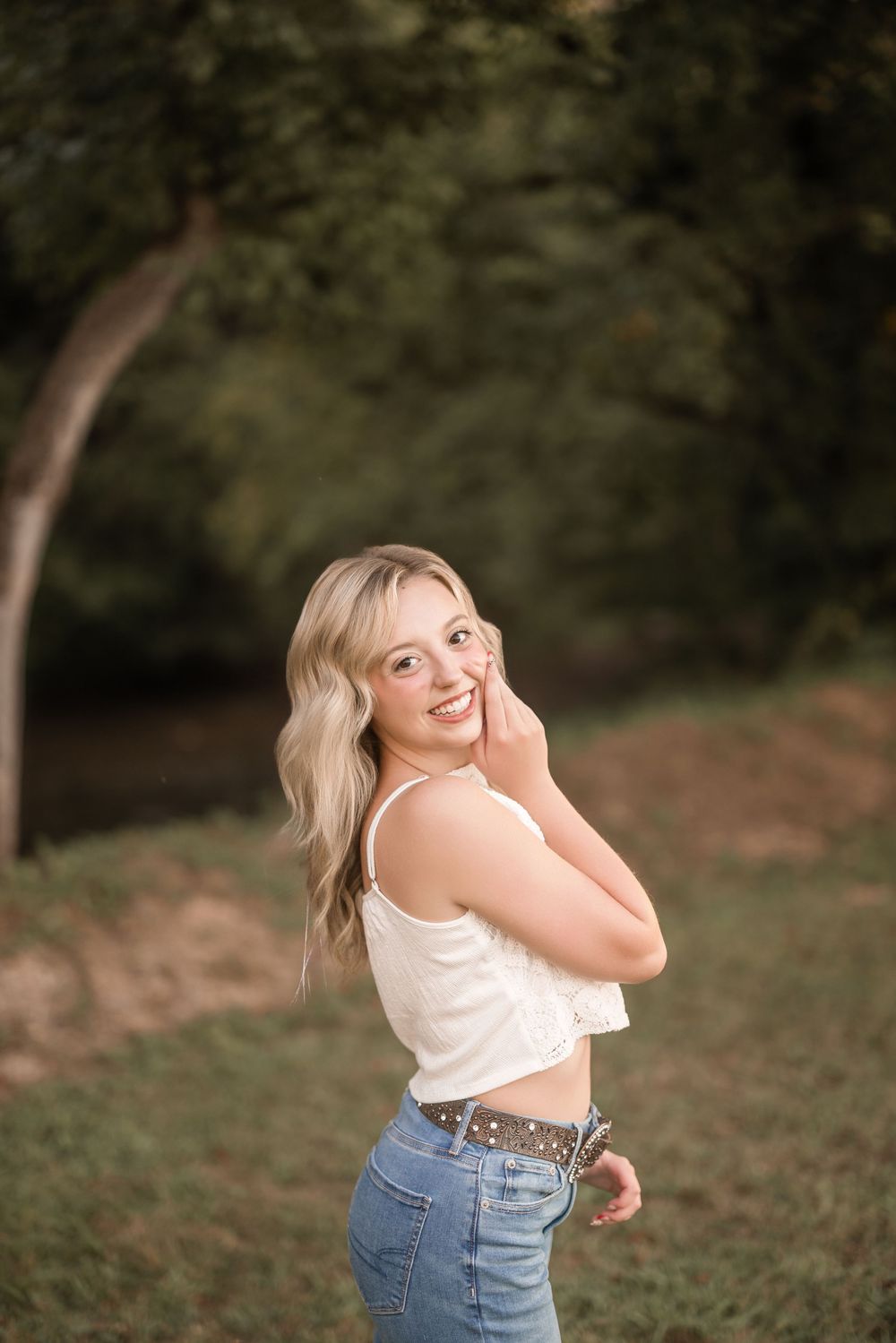 A person in a white crop top and jeans poses outdoors against a natural blurred background.