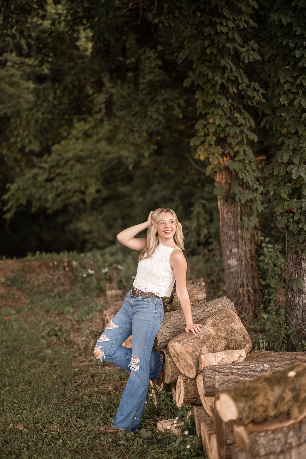 A summery scene showing someone in a white lace top and flared jeans posing casually by a woodpile in a forest setting.