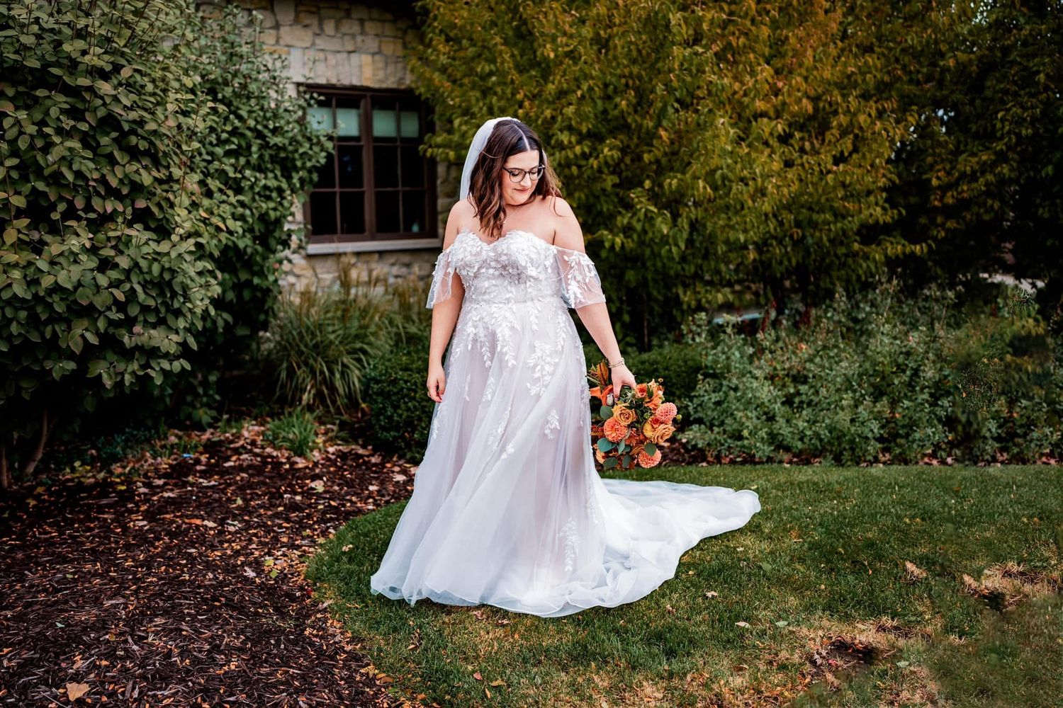A bride in a flowing white lace gown holds a vibrant autumn bouquet while standing in a garden setting.