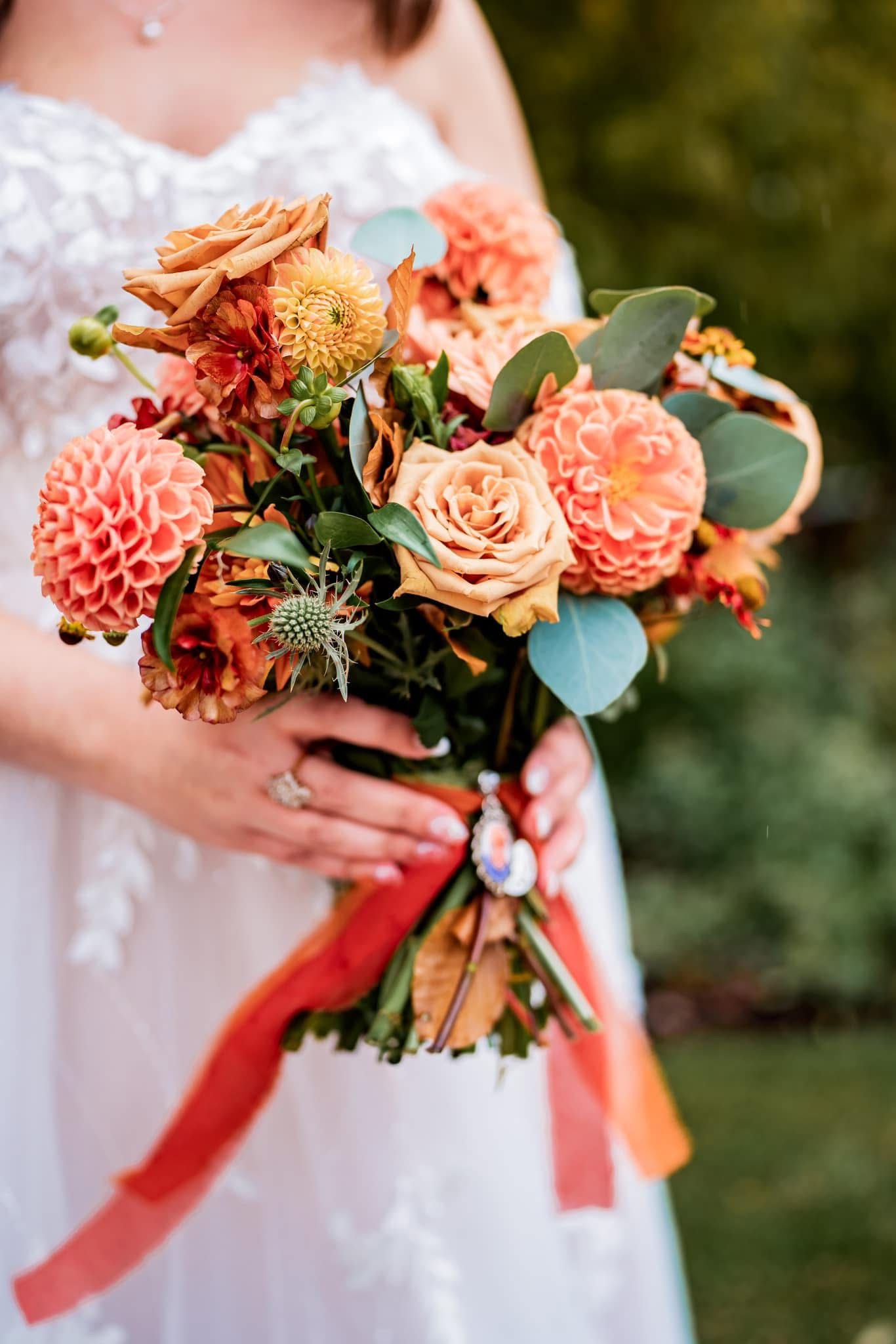 A coral and peach wedding bouquet with dahlias, roses, eucalyptus leaves, and flowing red ribbons held against a white dress.