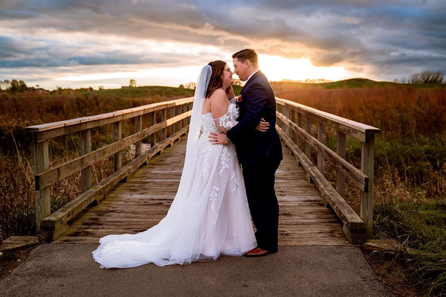 A couple in wedding attire embrace on a wooden boardwalk at sunset with dramatic clouds and golden light.