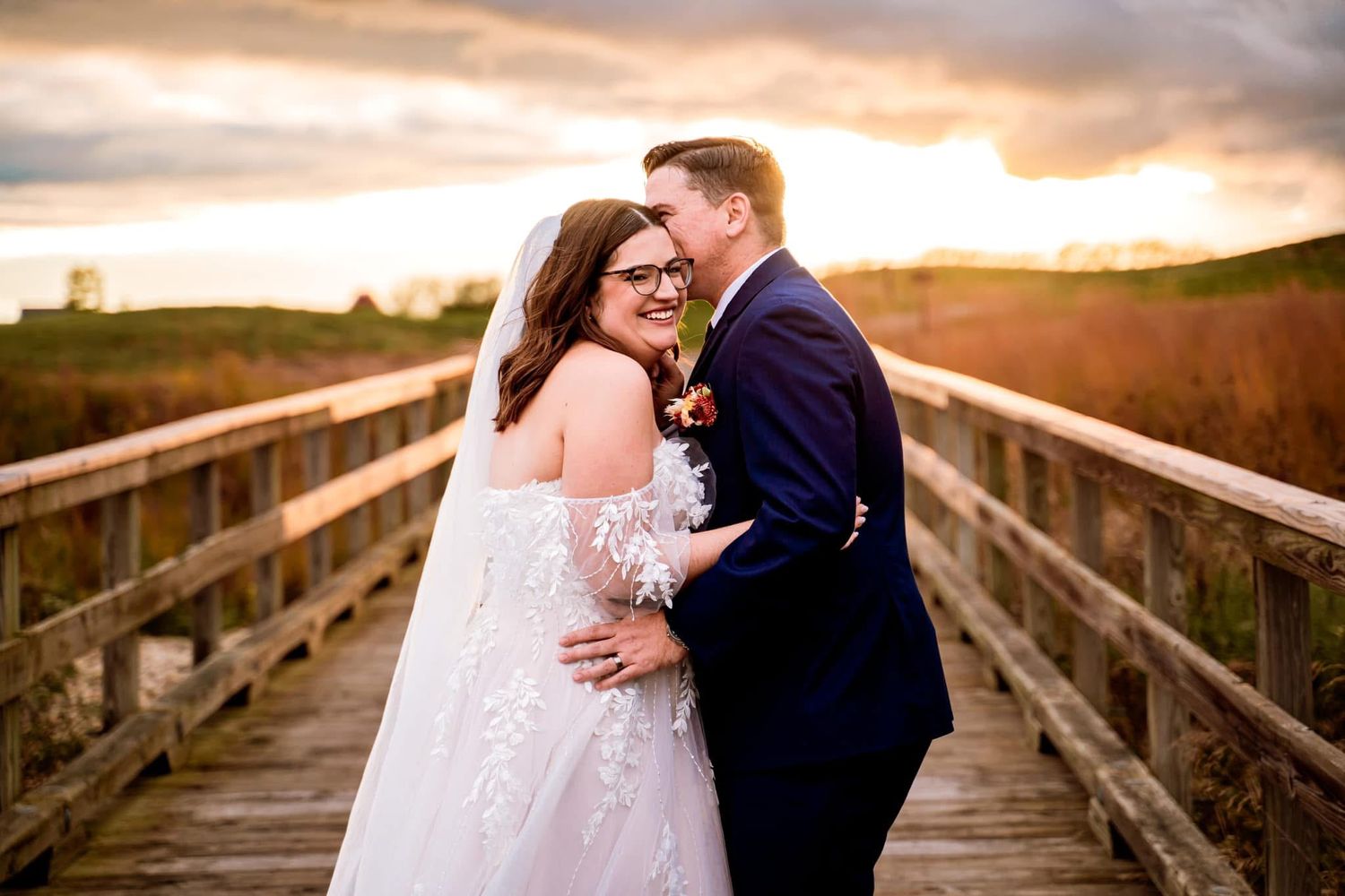 A couple embraces on a wooden boardwalk during sunset with golden light and cloudy sky in the background.