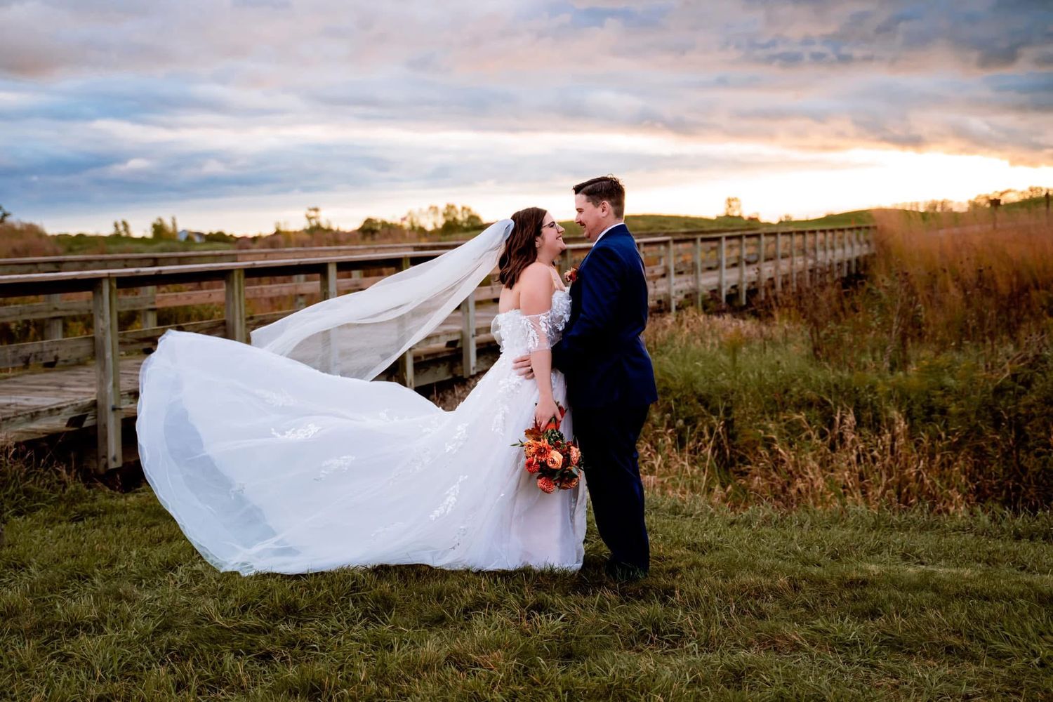 A couple poses on a wooden bridge at sunset while a flowing white wedding dress catches the breeze.