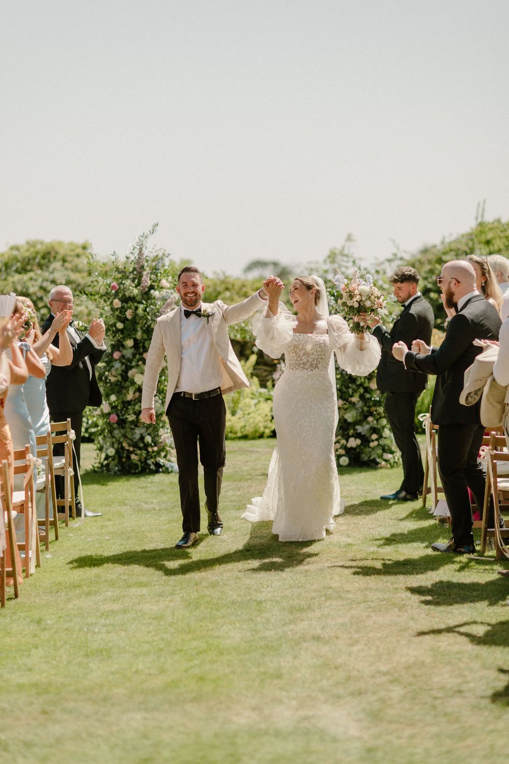 A joyful celebration on a grassy lawn as wedding guests cheer for a newly married couple walking down the aisle.