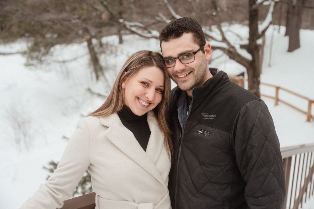 A couple stands close together smiling in a snowy winter landscape with evergreen trees.