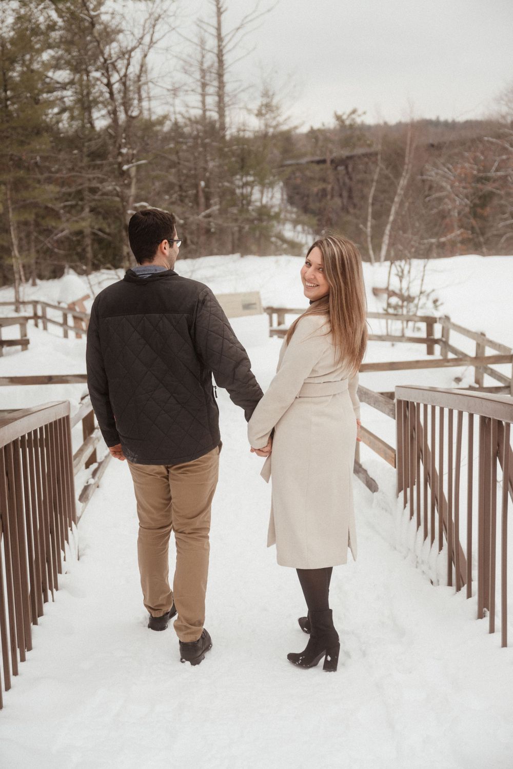 A couple walking hand in hand on a snow-covered wooden bridge in winter.
