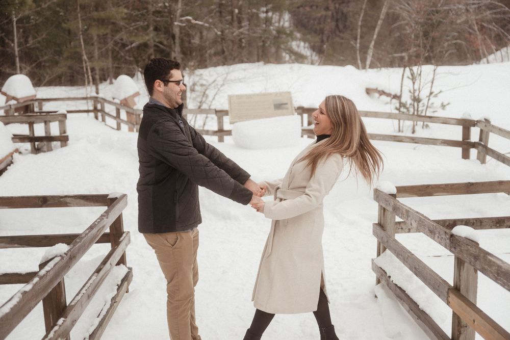 A playful couple holding hands and dancing on a snowy bridge in winter.