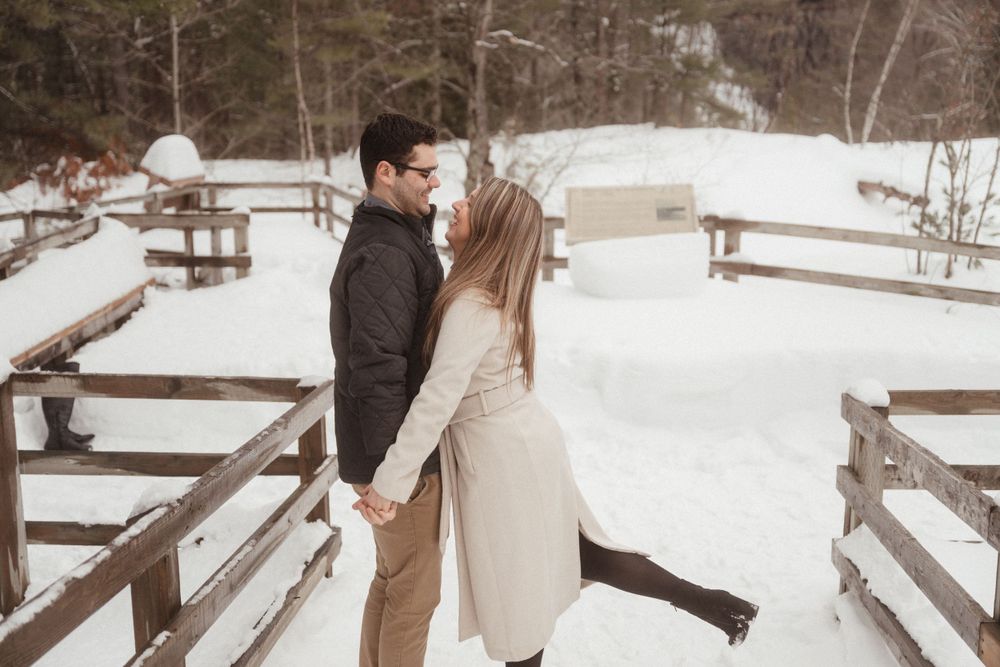 Romantic winter engagement photo of a couple embracing on a snowy wooden walkway with a bridge in the background.