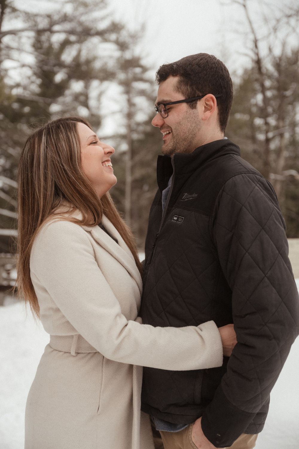 Joyful couple laughing together during winter photoshoot in warm coats against snowy landscape.