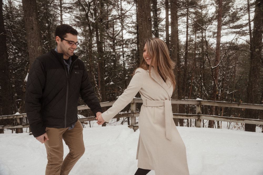 Playful couple dancing in the snow on wooden walkway surrounded by pine trees.