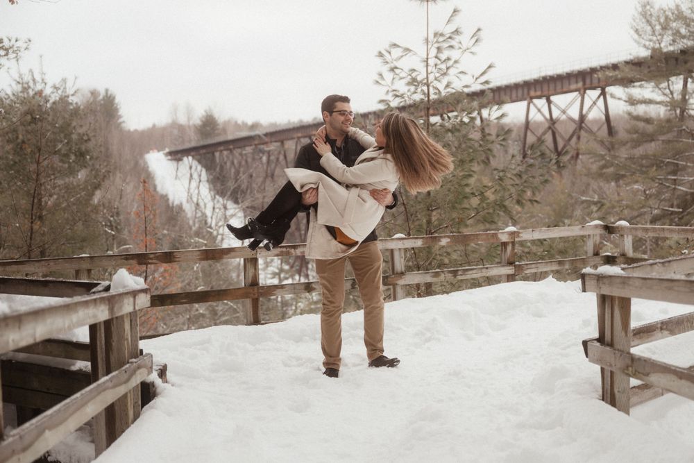 Romantic winter scene with couple in dramatic lift pose on snowy wooden bridge.