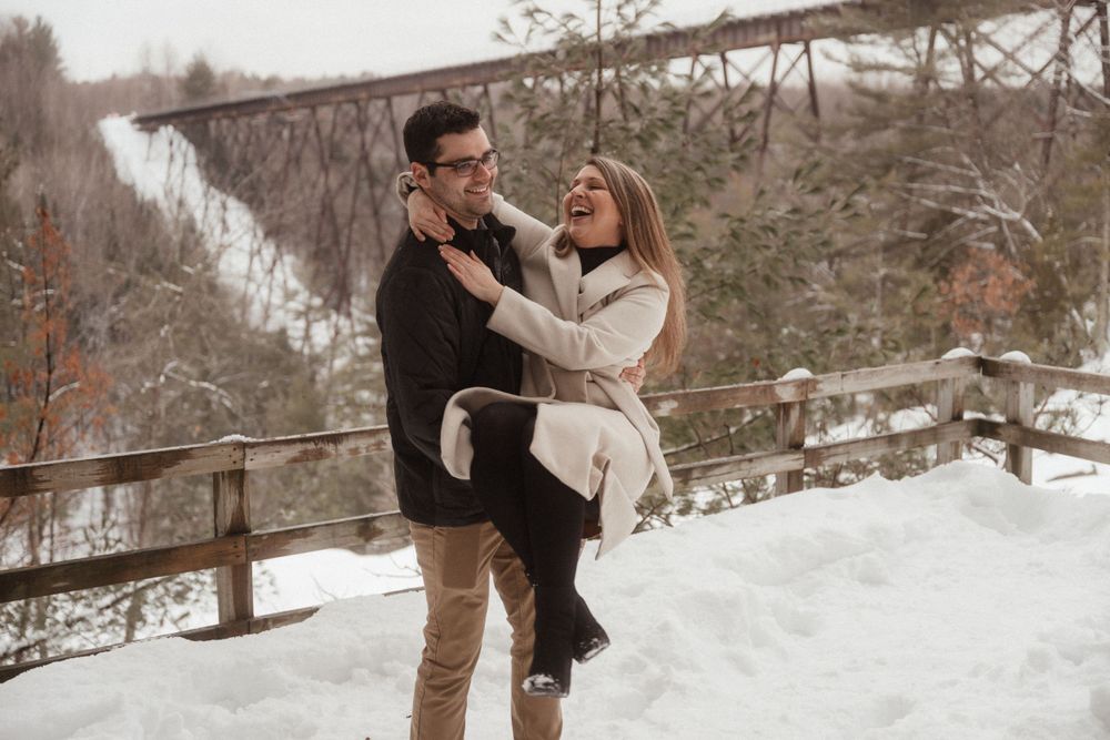 Playful winter photoshoot with couple in lifting pose against snowy bridge backdrop.