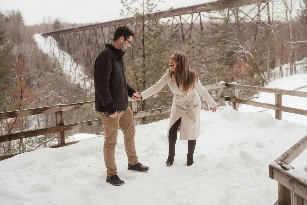 Couple playing and holding hands on wooden walkway during snowy winter photoshoot.