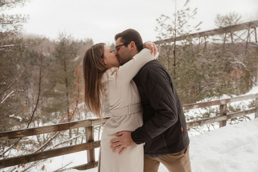 Intimate winter engagement photo with couple kissing on wooden walkway with bridge backdrop.