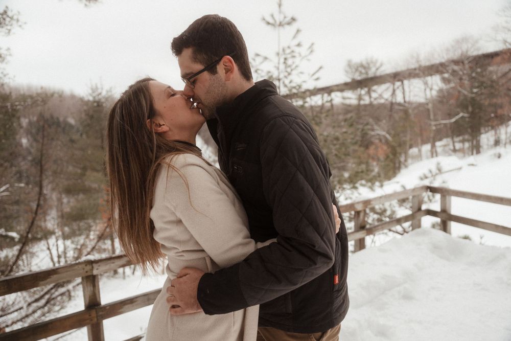 Romantic winter kiss between couple on snowy wooden walkway with rustic bridge in background.